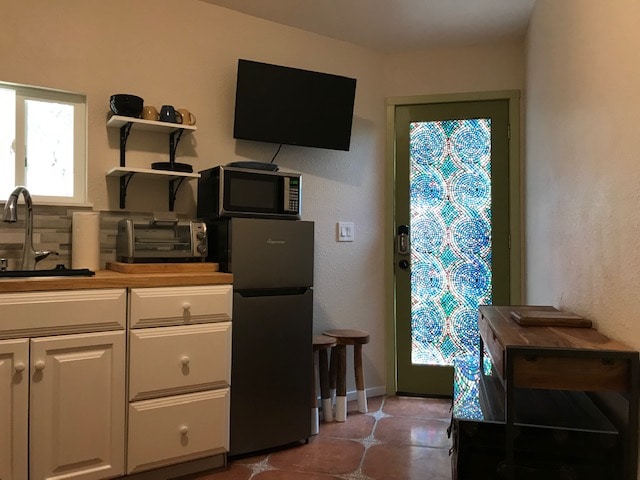 A bright kitchenette area is equipped with essential appliances, including a refrigerator, microwave, and toaster oven. Sturdy shelves hold dishware and a coffee maker. A wooden countertop offers additional workspace, while a vibrant glass door invites natural light into the space.