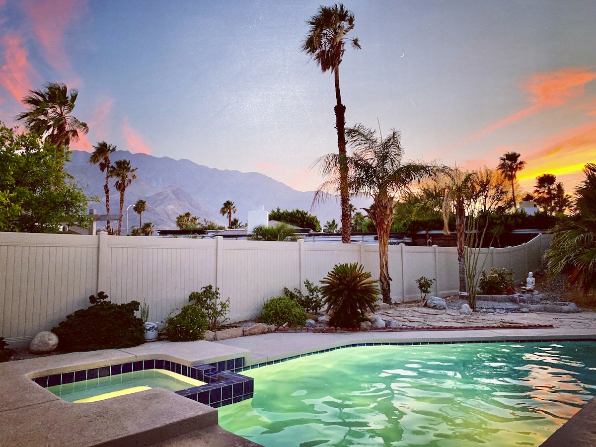 A private pool is surrounded by a peaceful patio, featuring lush greenery and palm trees. The mountains rise in the background, with a sunset sky painted in shades of orange and purple. The pool area offers a serene escape for relaxation.