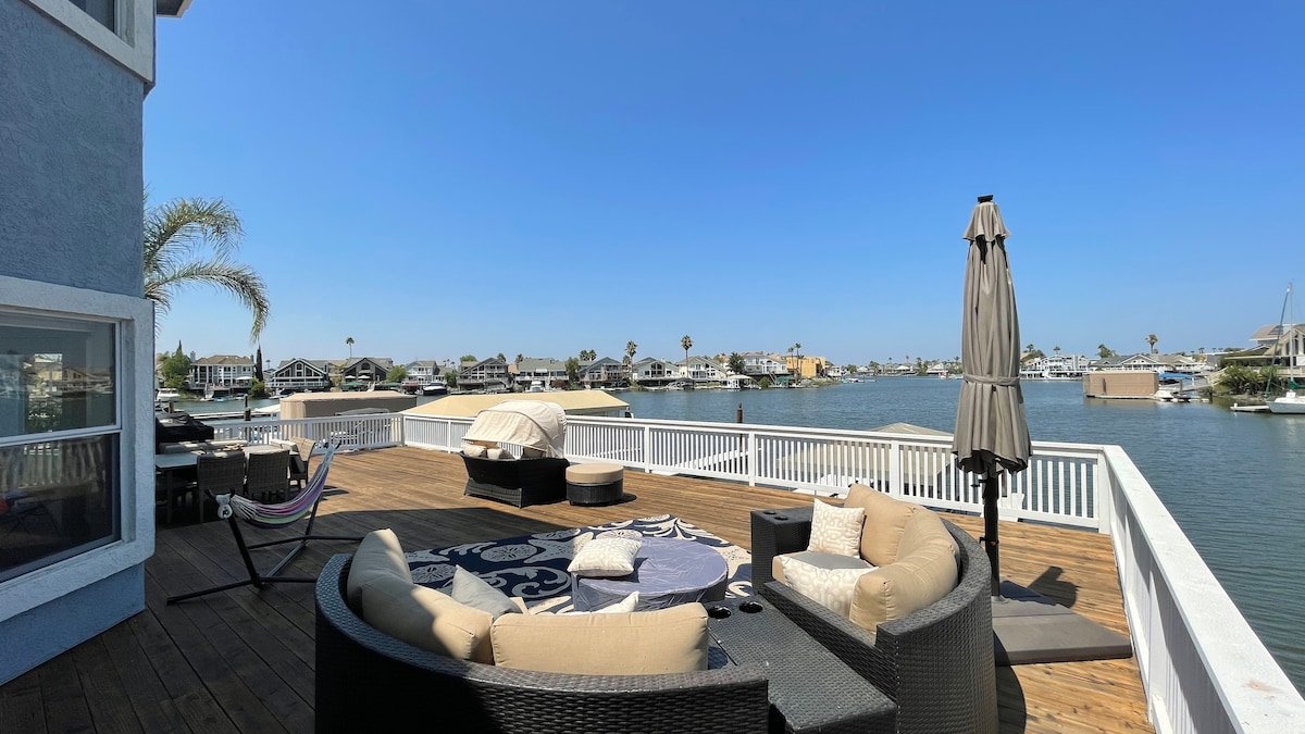 A wide wooden deck overlooks the waterfront, furnished with comfortable circular seating and an umbrella for shade. A hammock is positioned nearby, with views of surrounding homes and boats reflecting off the water under clear blue skies.