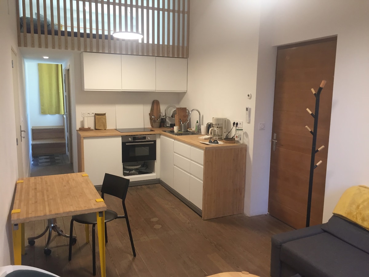 A well-organized kitchen and living area feature modern cabinetry and appliances. A wooden dining table is complemented by a black chair. Natural light enters through a window, and a staircase leads to the mezzanine. Walls are finished in light tones, contributing to a spacious feel.