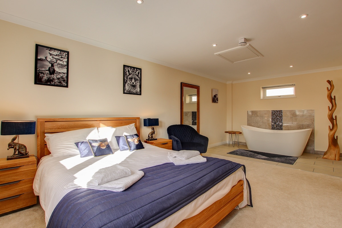 A spacious bedroom includes a comfortable bed with crisp linens and decorative pillows. Natural light brightens the room, enhancing the elegant slipper bath and the stylish chair nearby. Framed artwork adorns the walls, contributing to a serene ambiance.
