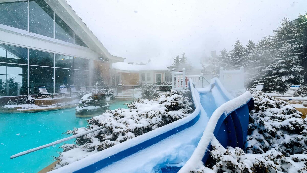 A winter scene captures a blue water slide surrounded by snow-covered landscaping. The pool is visible in the background, steam rising in the chilly air. Large windows of the adjacent building showcase the indoor space, while snowflakes gently fall from the sky.