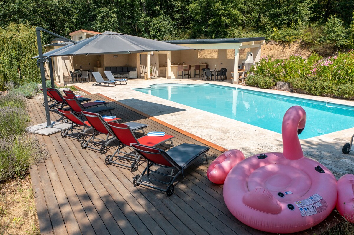 A swimming pool is framed by sun loungers on a wooden deck. An outdoor kitchen and dining area are shaded by a canopy, with lush greenery in the background. A pink inflatable flamingo floats in the water, adding a vibrant touch to the scene.