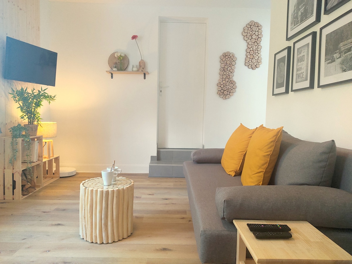 A comfortable living area features a gray sofa adorned with bright yellow pillows, offering a welcoming touch. A wooden coffee table, resembling logs, is placed in front, while a television is mounted on the wall. Soft lighting enhances the space's cozy ambiance.