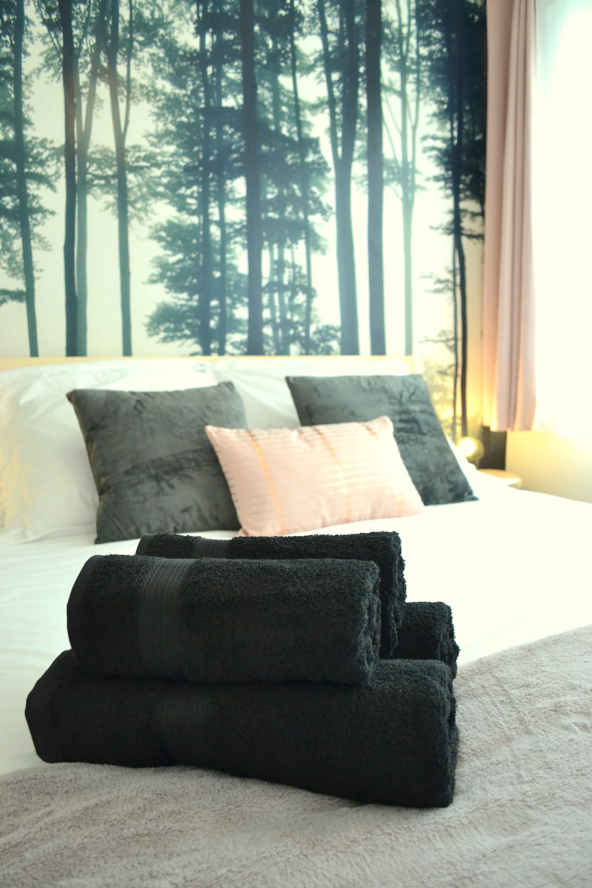 A well-made bed features soft white linens and decorative pillows, framed by a calming forest-themed wall mural. Neatly rolled black towels are placed on the bed, adding a touch of contrast against the light bedding.