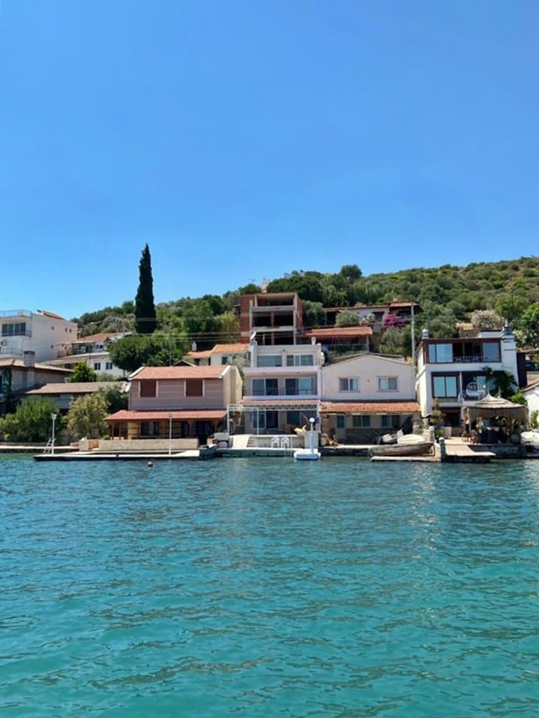 A row of multi-storied homes is observed along the waterfront, showcasing various architectural styles. Lush greenery and trees are positioned on the hillside behind the houses. The calm, clear water reflects the buildings under a bright blue sky, creating a serene coastal view.