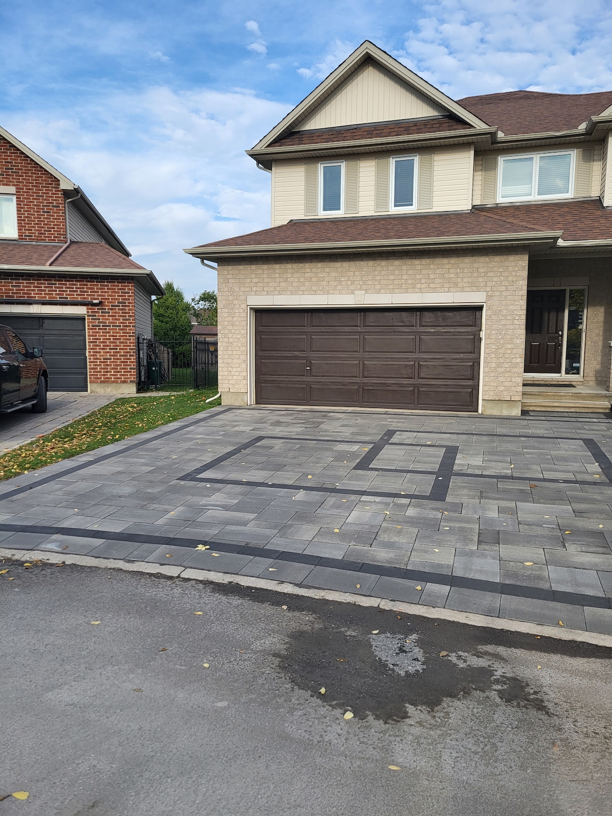 A driveway with patterned paving leads to a two-car garage featuring a brown door. The beige brick facade of the house complements the light gray paving stones. Nearby, a well-maintained lawn provides a touch of greenery against the structured surfaces.