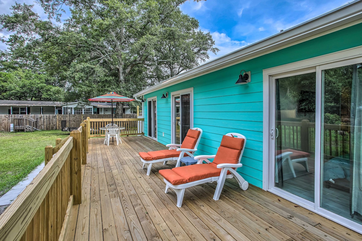 A spacious deck features two lounge chairs with orange cushions, inviting relaxation under the open sky. The bright blue exterior of the cottage is visible, along with a shaded dining area under an umbrella. Lush greenery surrounds the area, enhancing outdoor enjoyment.