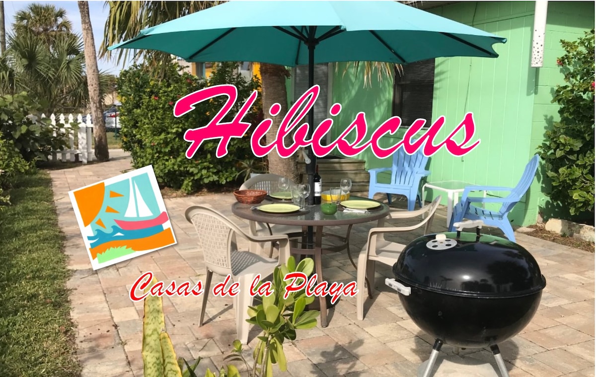 An outdoor dining area is featured with a round table surrounded by four chairs. An umbrella provides shade, and a charcoal grill is positioned nearby. Vibrant tabletop decor is visible amidst the tropical landscaping, contributing to a relaxed atmosphere.
