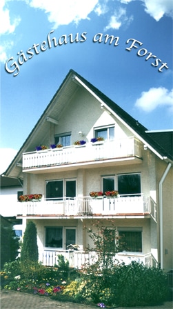 The exterior of the Gästehaus am Forst is showcased, featuring a three-story building with white walls and balconies adorned with flower boxes. A well-tended garden and pathway lead up to the entrance, under a clear blue sky with a scattering of clouds.