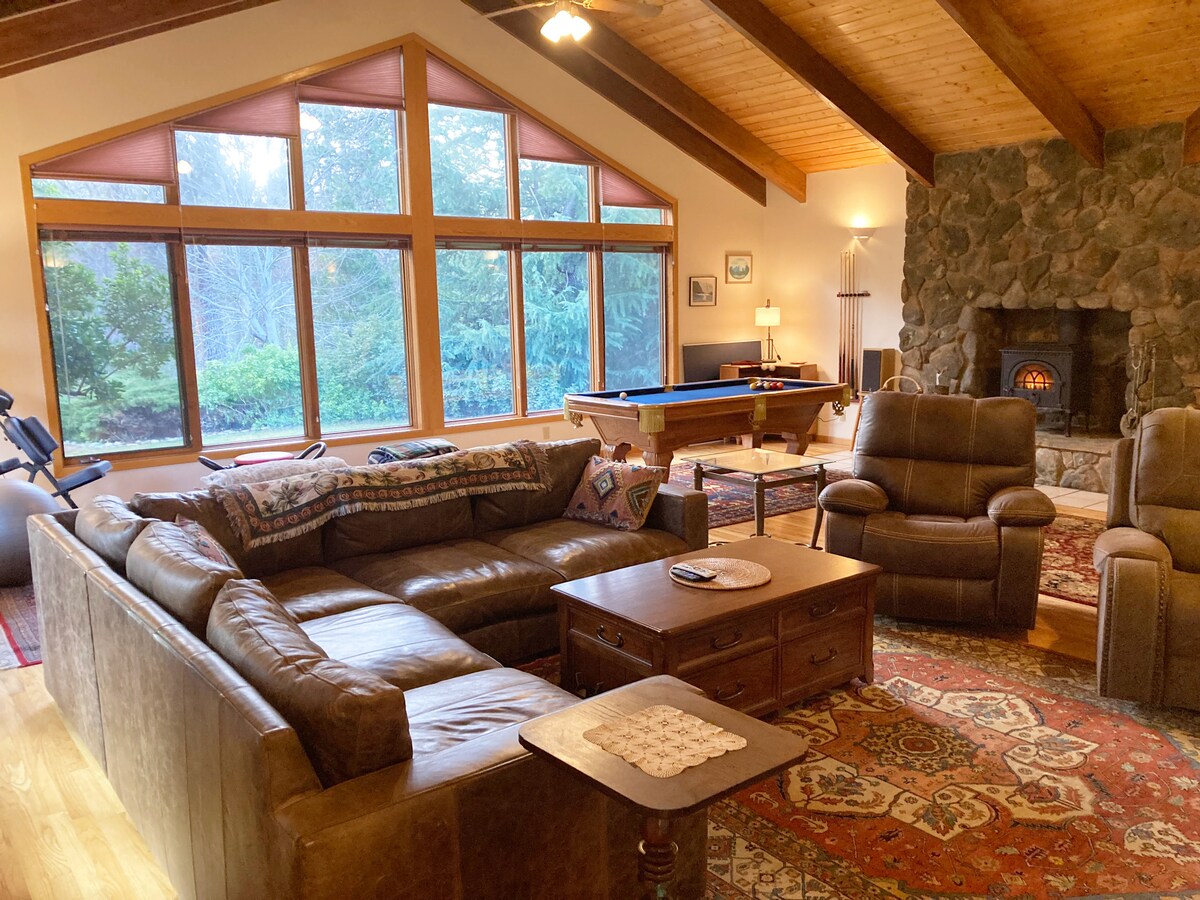 A spacious living area features a comfortable leather sectional sofa, positioned near a stone fireplace. Large windows allow natural light to fill the room, showcasing views of the surrounding greenery. A pool table is visible in the background, adding to the entertainment options.