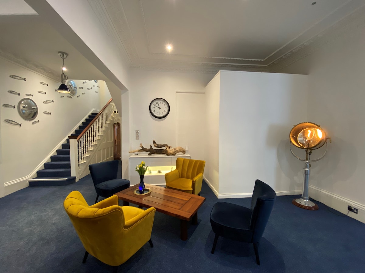 A welcoming lounge area is presented with a wooden coffee table surrounded by yellow and navy armchairs. A large wall clock is mounted above, while decorative fish artwork is visible on the walls. A stylish lamp enhances the space, which features blue carpeting.