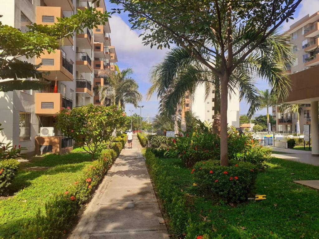 A walkway lined with lush greenery and flowering plants leads through the residential complex. Tall palm trees provide shade along the paved path, with buildings visible in the background. The area is well-maintained, offering a peaceful environment for leisurely strolls.