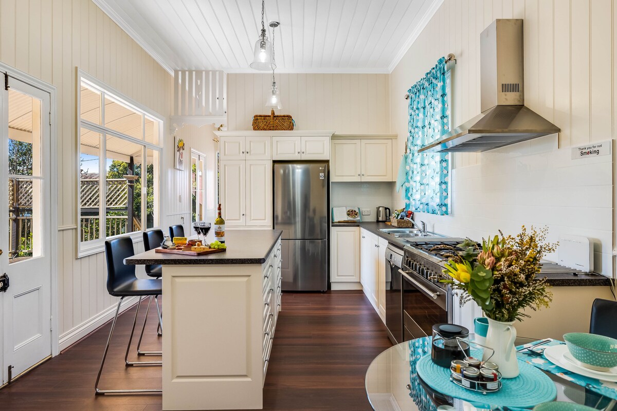 A modern kitchen features a central island with seating and a spacious layout. Natural light fills the area through large windows, complemented by light-colored cabinetry. A dining table is set for guests, showcasing dishes and a floral arrangement, while appliances are neatly arranged along the wall.