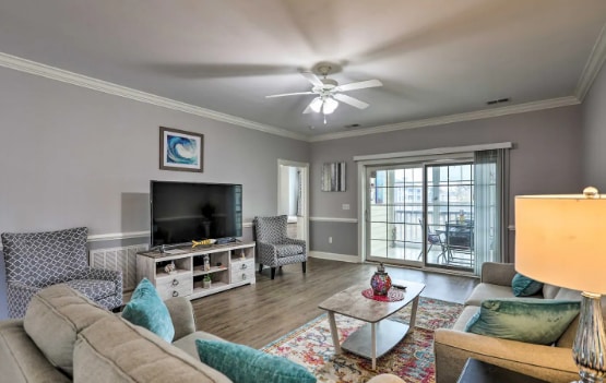The living area is furnished with a large beige couch, two patterned chairs, and a coffee table. A TV is placed on a light wood console. Natural light floods the room through sliding glass doors leading to a patio with seating.