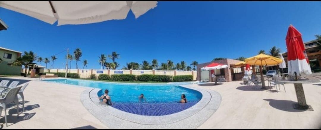 A spacious outdoor pool area is surrounded by palm trees, with sun loungers positioned under umbrellas. Families can be seen enjoying the water. Colorful umbrellas provide shade, and the pool's blue water reflects the clear sky. A relaxing atmosphere is highlighted by the well-maintained surroundings.