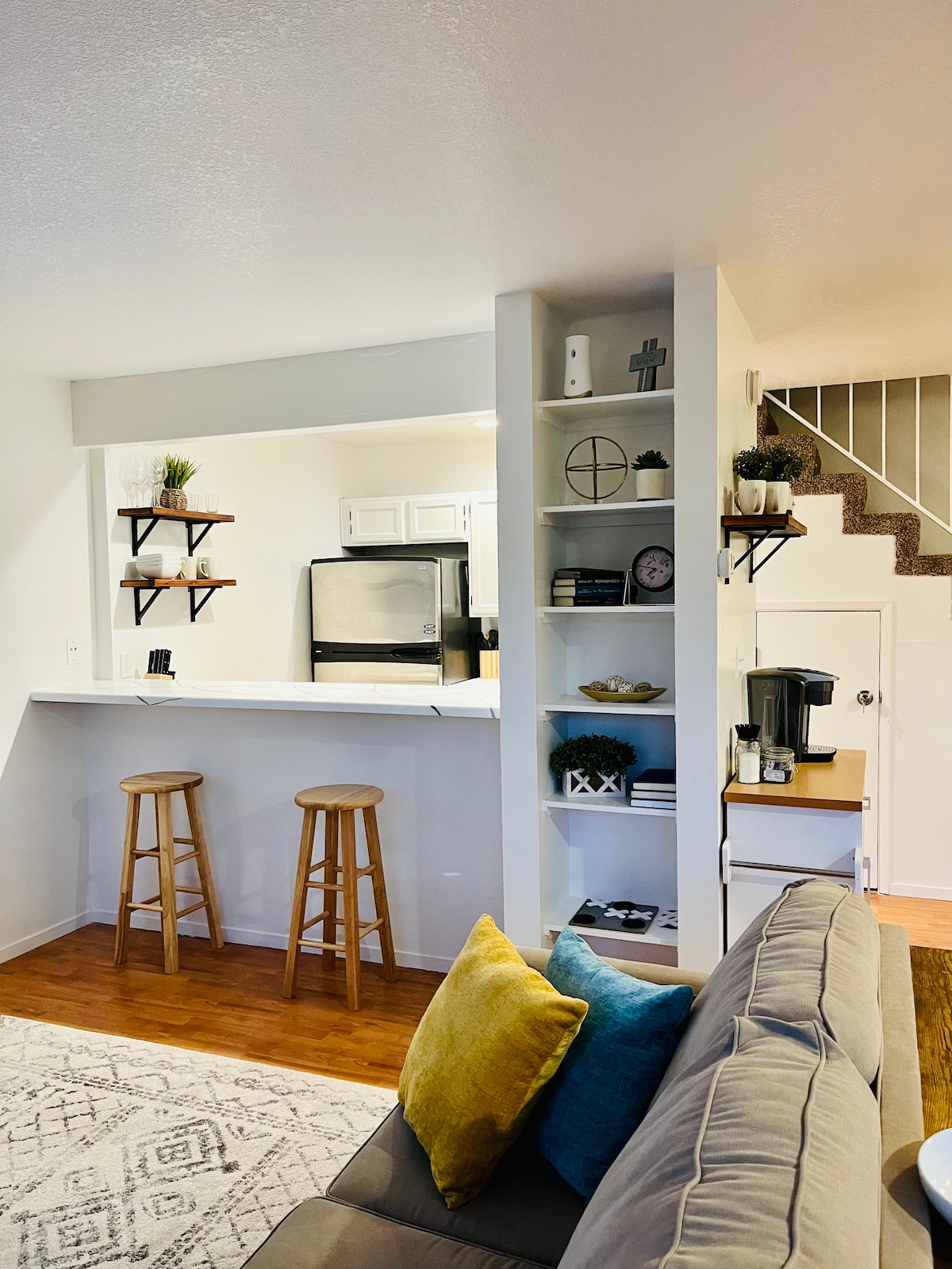 The living area features a comfortable couch with vibrant pillows. A sleek, open kitchen area is visible in the background, framed by modern shelving. Two wooden stools are positioned at the counter, and a staircase leads to the upper level.