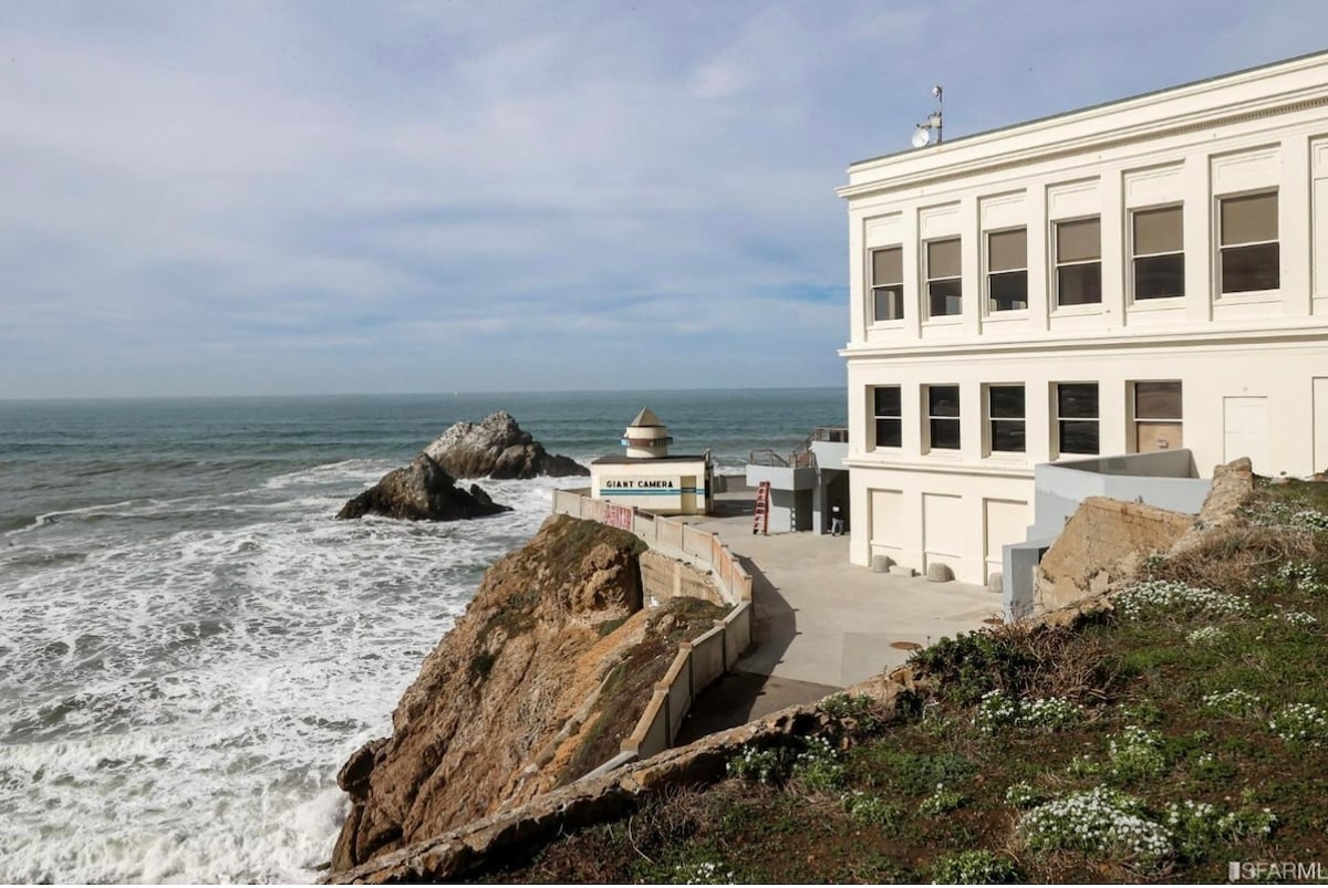 The image shows a coastal structure perched on a rocky shoreline, with waves crashing against the cliffs below. A viewpoint is visible adjacent to the building, highlighting the proximity to the ocean. The building’s exterior features large windows reflecting the scenic coastal view.