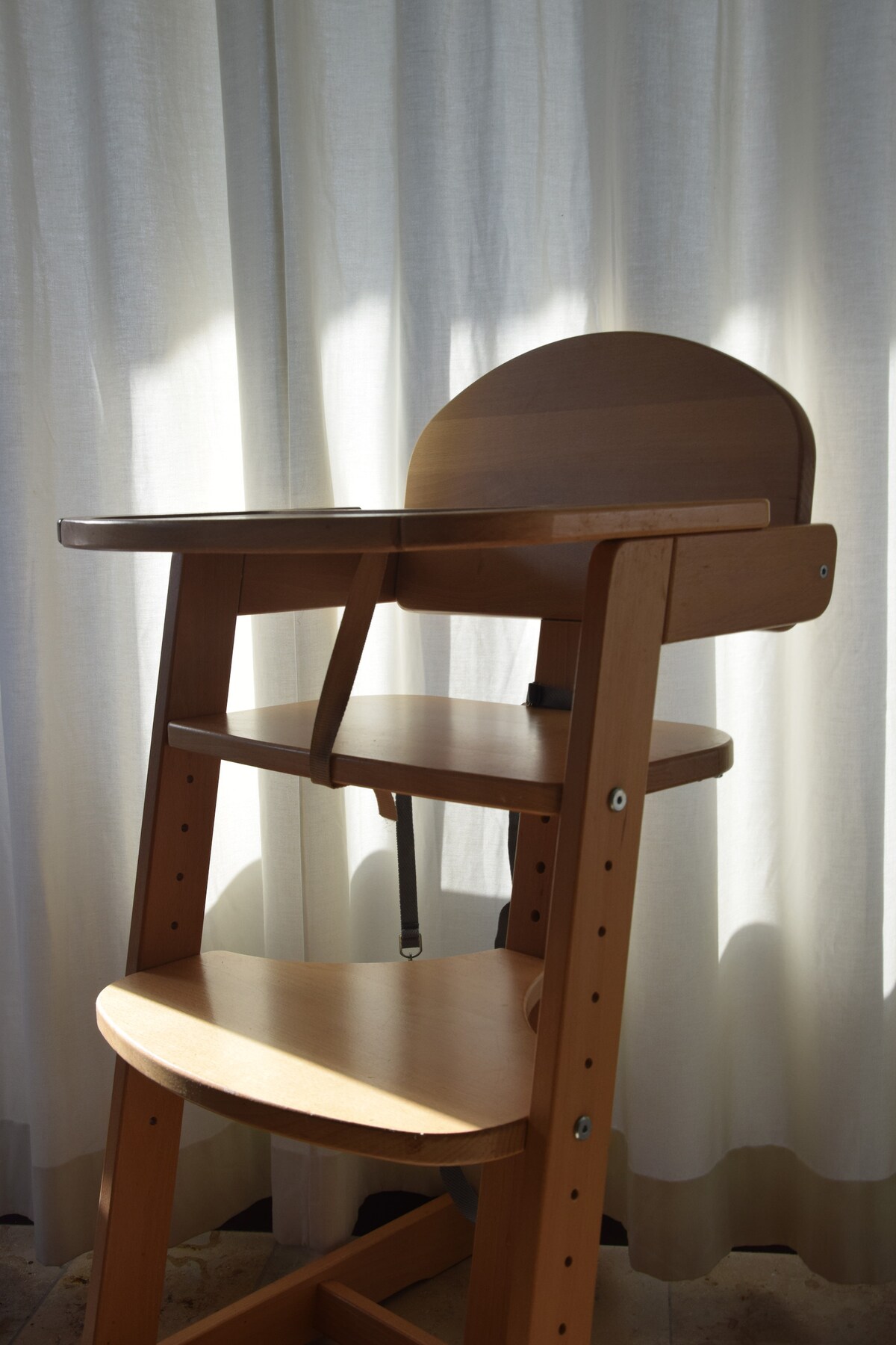 A wooden high chair is positioned near a softly lit window, which is framed by sheer curtains. The chair features a spacious seat with a supportive backrest and a tray for meals, designed for comfort and safety.
