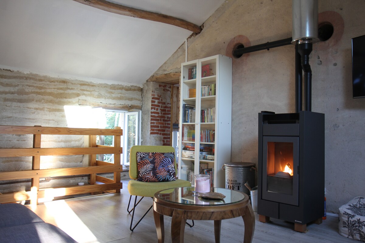 A cozy living area features a modern stove providing warmth. A circular table sits centrally, surrounded by two chairs, one with a patterned cushion. Natural light floods in through a large window, highlighting the rustic charm of the exposed brick and wooden beams.