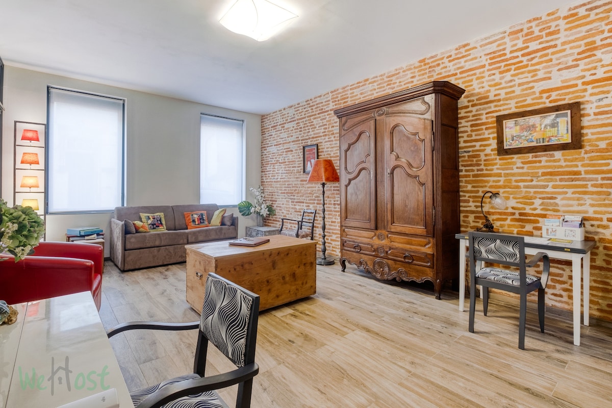 A spacious living area features exposed brick walls and warm wood flooring. A comfortable sofa sits adjacent to a wooden coffee table. An antique armoire adds character, while modern chairs provide additional seating. Soft light filters through the windows, enhancing the room's inviting ambiance.