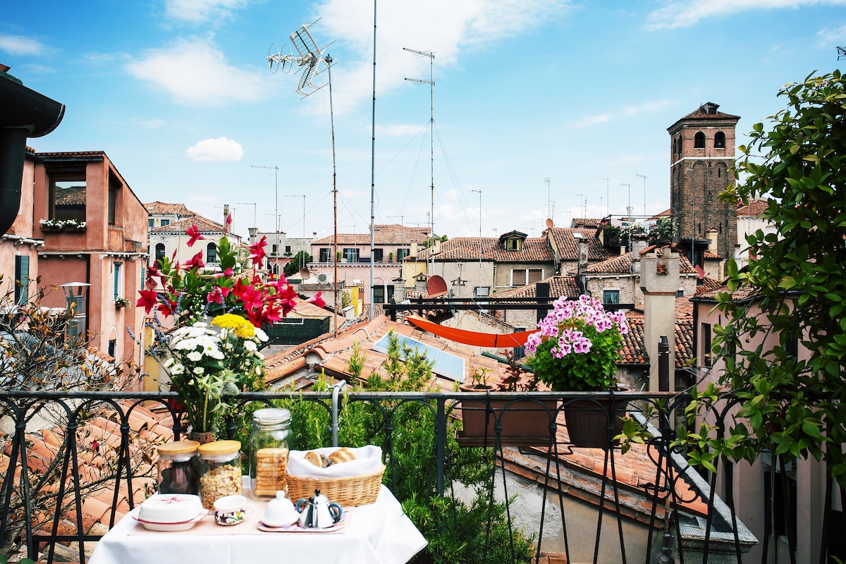 A balcony table is set with a white tablecloth, featuring a basket, jars, and breakfast items. Colorful flowers in pots enhance the view of terracotta rooftops and distant buildings under a clear blue sky. Television antennas and a bell tower are visible in the background.