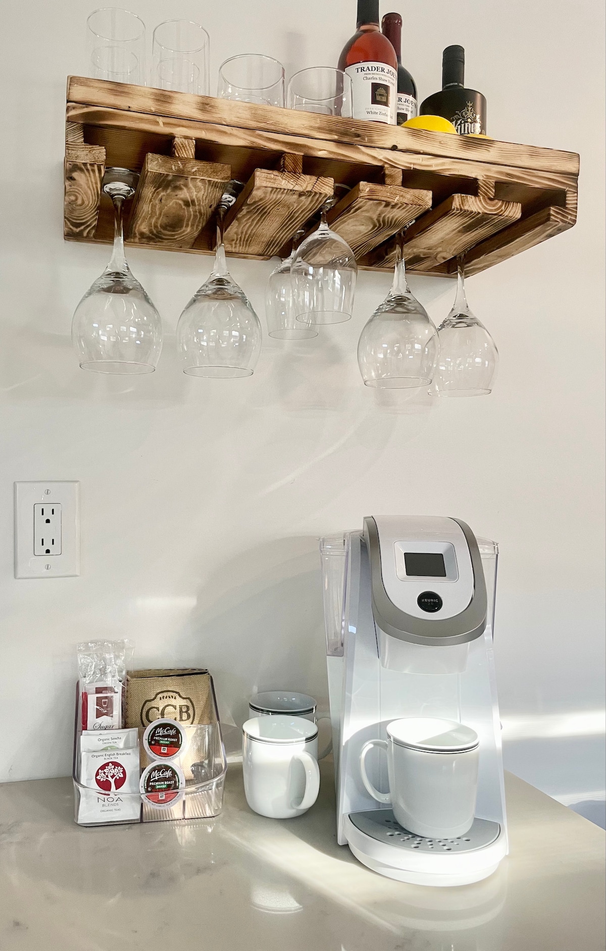 A coffee station features a modern brewing machine positioned next to a white ceramic mug. Above, a wooden shelf holds several wine glasses and bottles, along with fresh lemons, contributing to a well-organized space. The countertop exhibits a selection of coffee supplies.