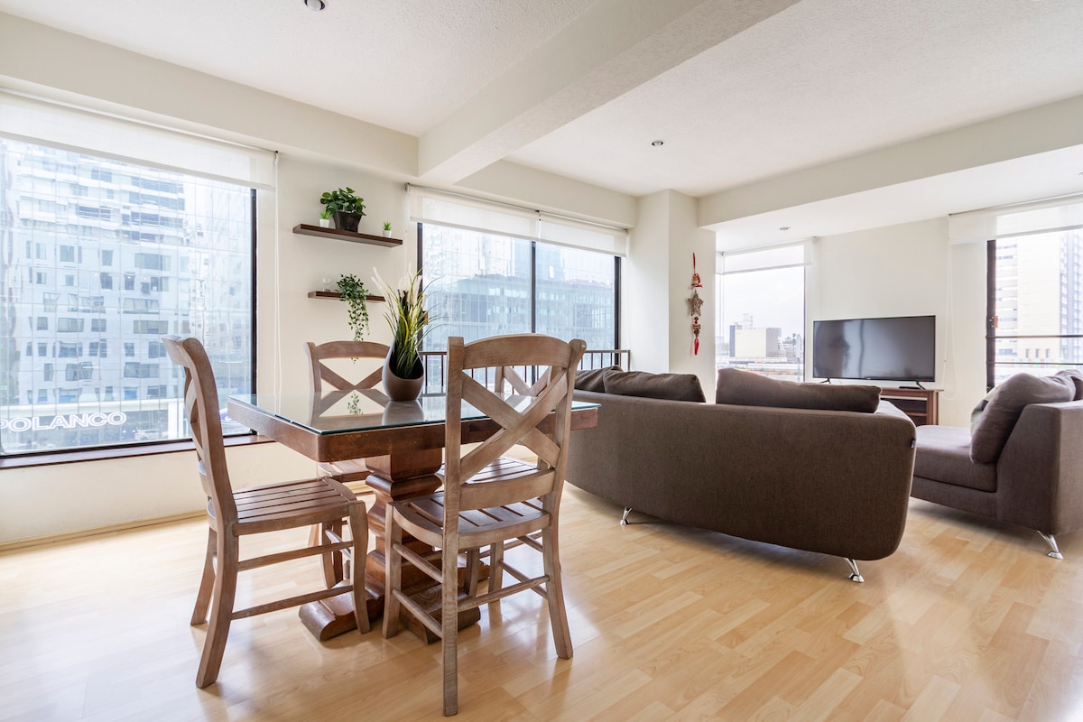 A well-lit living area is presented with large windows offering city views. Light wood flooring complements a wooden dining table surrounded by four chairs. A cozy sofa faces a media unit, creating a comfortable space for relaxation and gatherings.