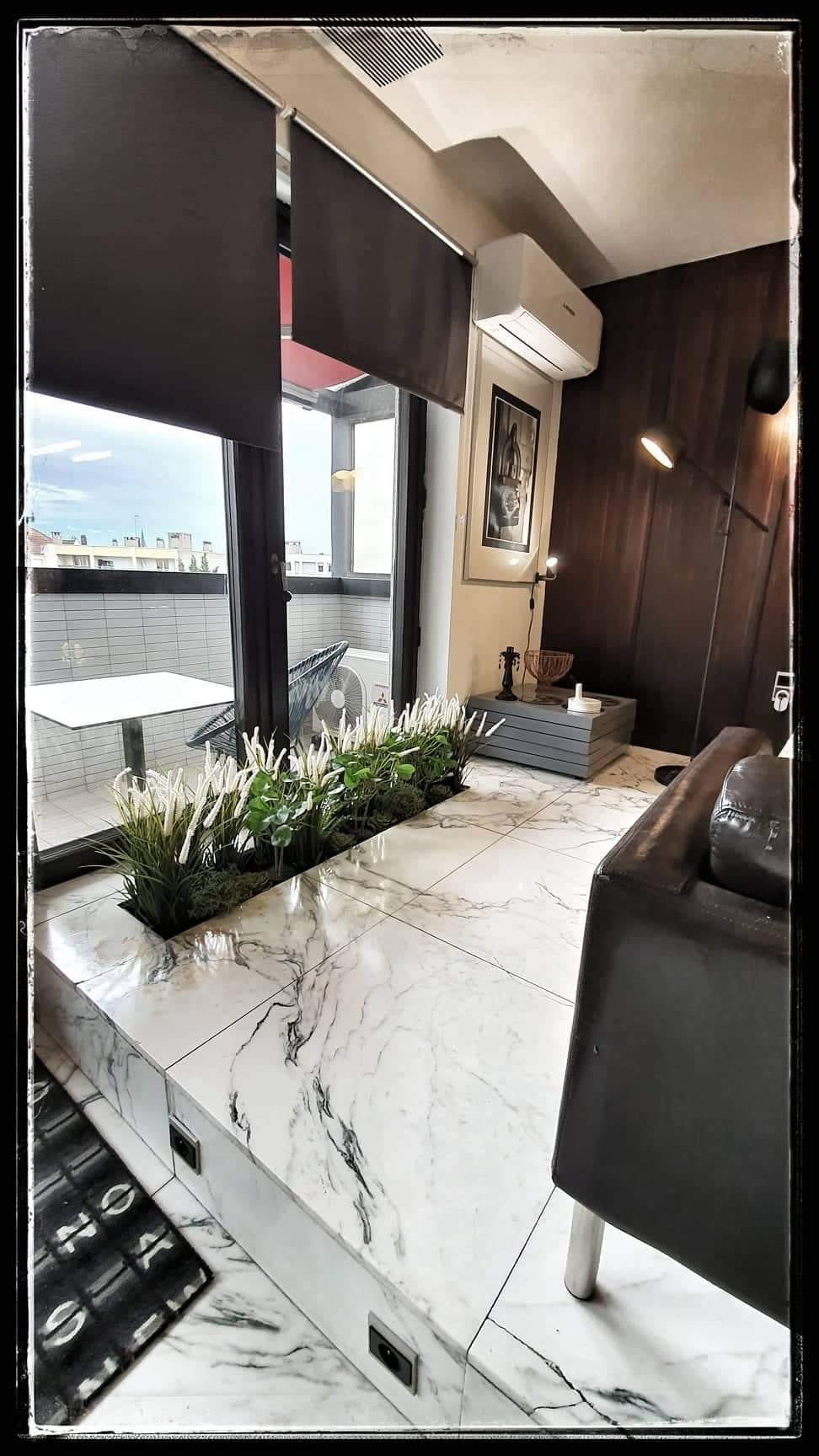 A stylish living area features a marble floor and large windows, offering natural light and views of a covered balcony. Indoor plants add a touch of greenery, while a modern chair and lighting create a cozy, inviting space.