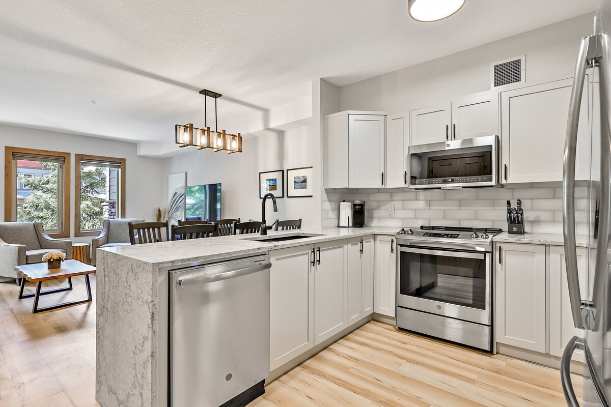 A modern kitchen features sleek cabinetry and a spacious countertop, complemented by stainless steel appliances. The area includes a dining space with dark wooden chairs and a light wood table. Natural light fills the room through large windows, enhancing the overall brightness.