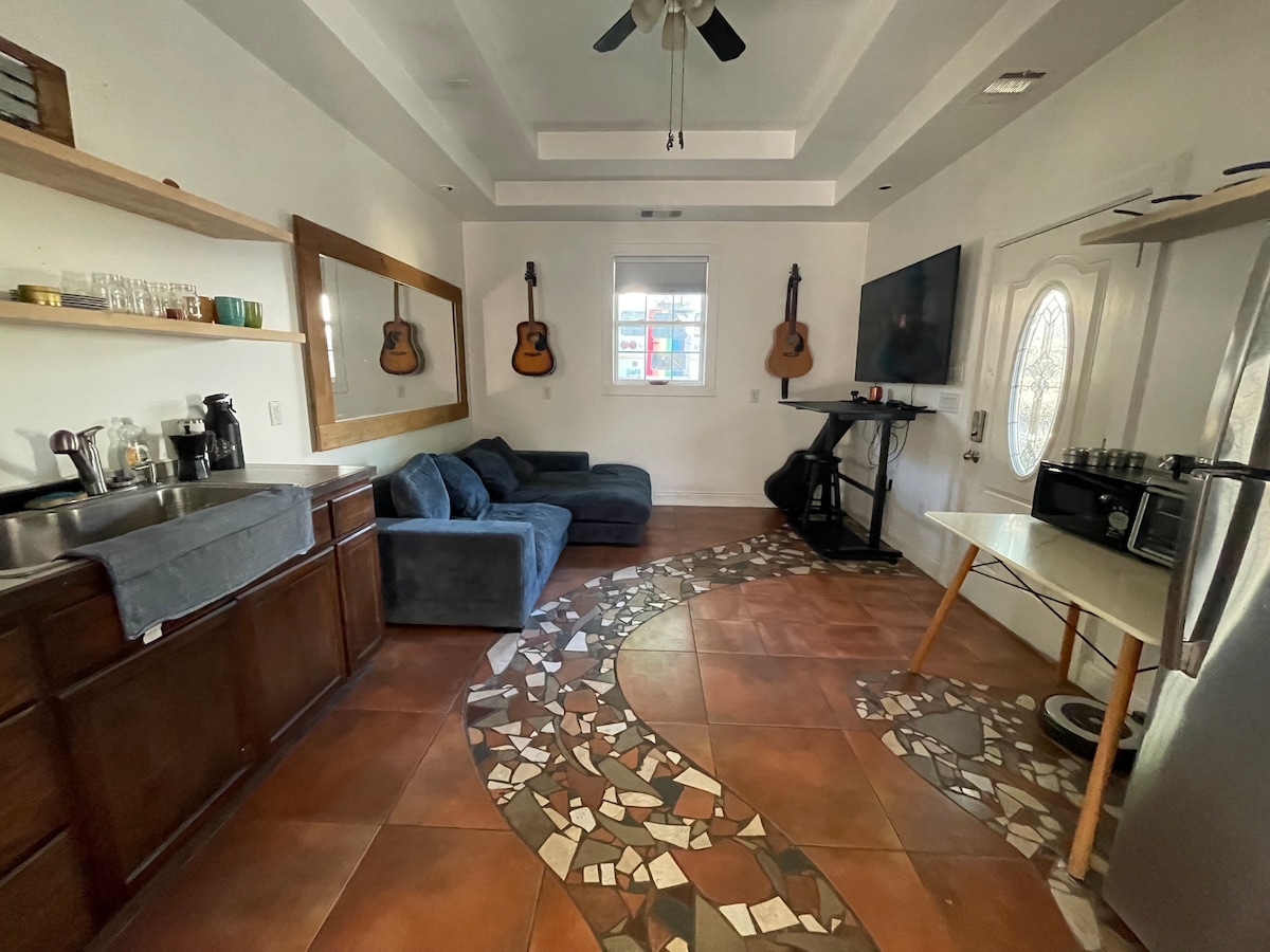 An inviting living area is showcased with a plush sectional sofa and decorative guitars mounted on the walls. A sleek television is positioned above a functional workspace. Light reflects off the unique patterned tile flooring, enhancing the contemporary and open layout.