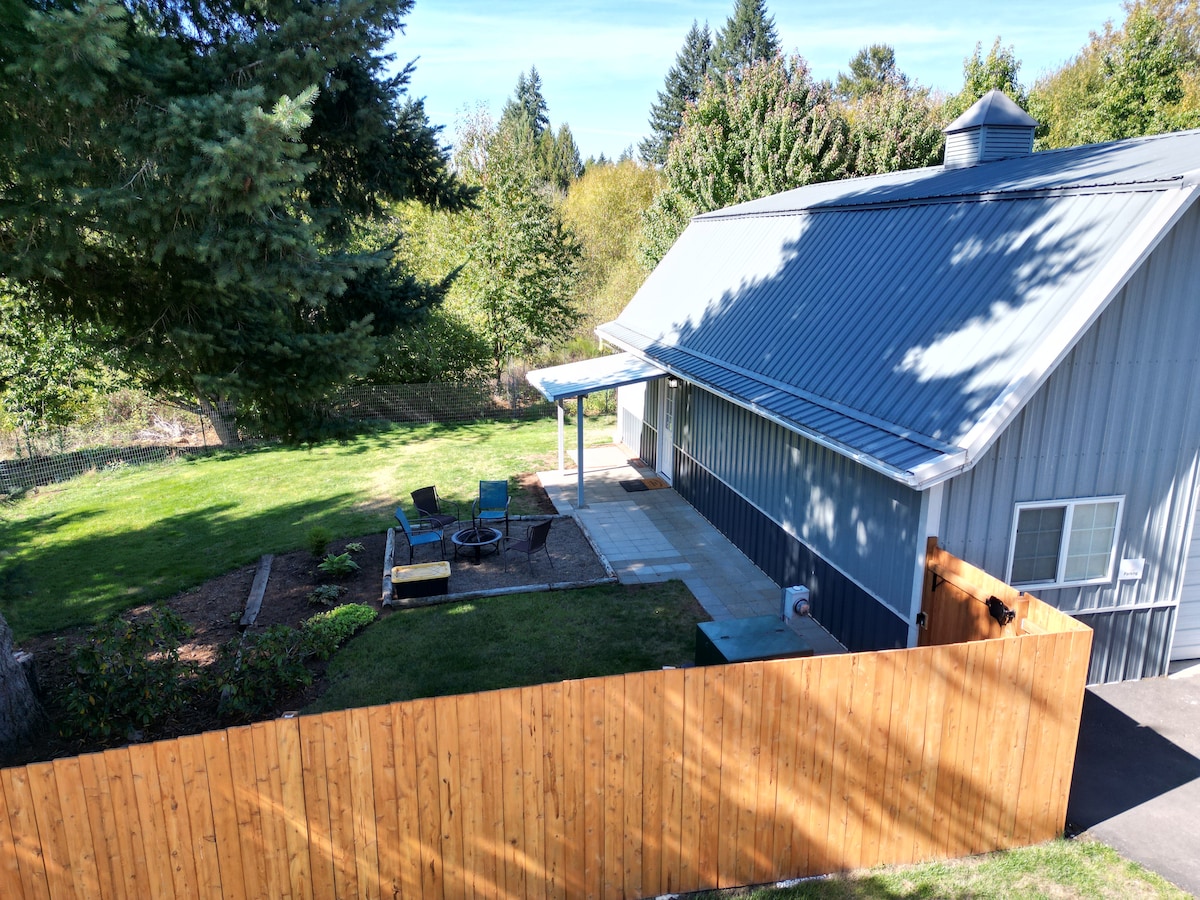An aerial view captures a fenced outdoor area surrounding a medium-sized dwelling, featuring green grass and a small patio. A fire pit with seating is visible in the yard, encompassed by trees and shrubs, promoting a sense of privacy and connection to nature.