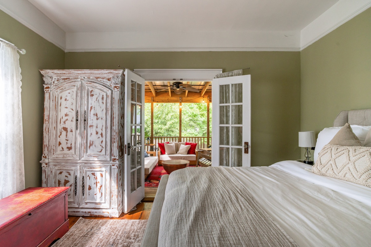 A cozy bedroom is showcased, featuring a spacious bed dressed in neutral linens. Double doors open to a screened-in porch, allowing natural light to flow in. A vintage armoire adds character, and soft green walls create a calming atmosphere. Brown wood floors complete the inviting space.