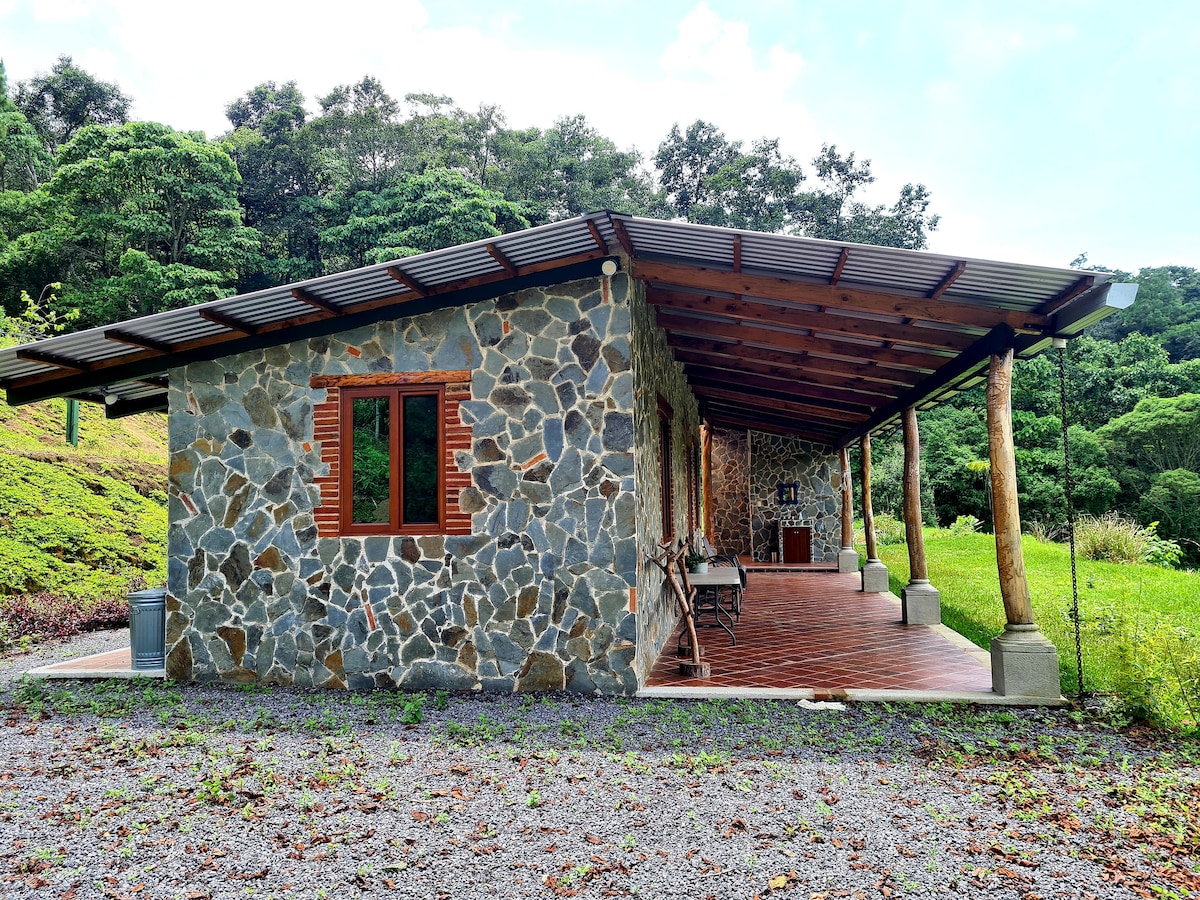 A rustic stone house is nestled among lush greenery, featuring a spacious covered porch with wooden beams. Large windows provide natural light, and wooden furniture is arranged on the porch. The surrounding landscape showcases vibrant foliage, enhancing the serene atmosphere of the setting.
