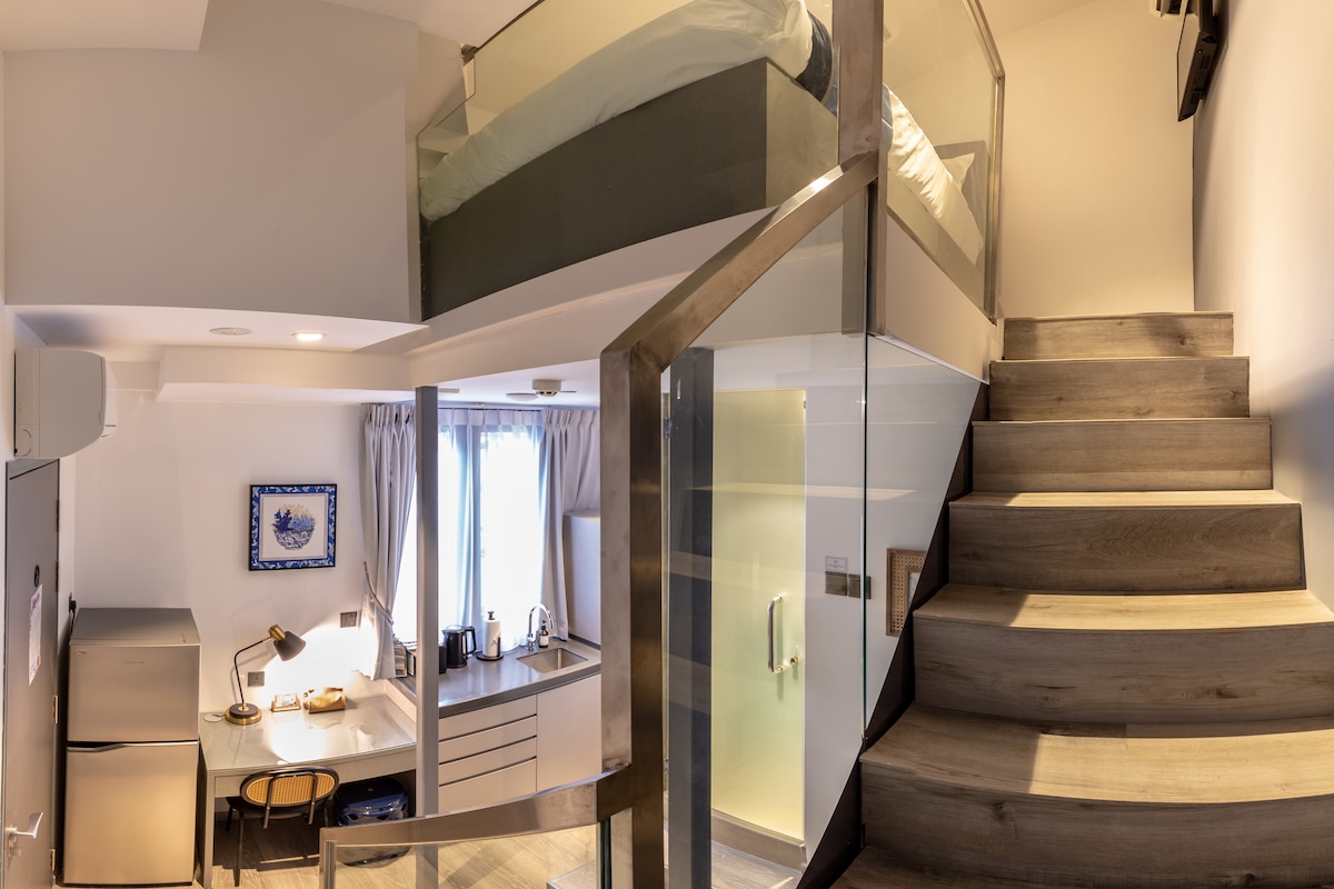A modern loft apartment is depicted, showcasing a clear view of the upper sleeping area above a cozy dining and kitchen space. Sturdy wooden stairs lead up, with glass railings providing safety and an open feel. Natural light floods the room through large windows.
