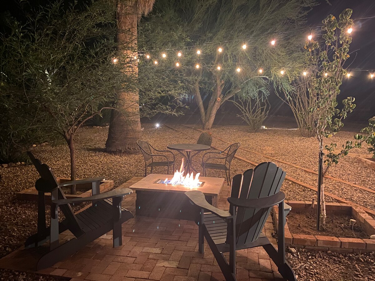 An outdoor seating area features four Adirondack chairs arranged around a fire pit. Strung lights illuminate the space, while palm trees and desert landscaping create a serene backdrop, inviting relaxation in the evening ambiance.