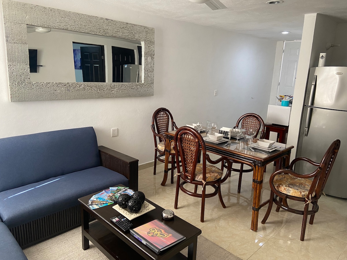 A comfortable seating area is complemented by a dining table for four, set with plates and glasses. The spacious kitchen is visible in the background, with a refrigerator and modern finishes. A large mirror reflects light, enhancing the open feel of the space.