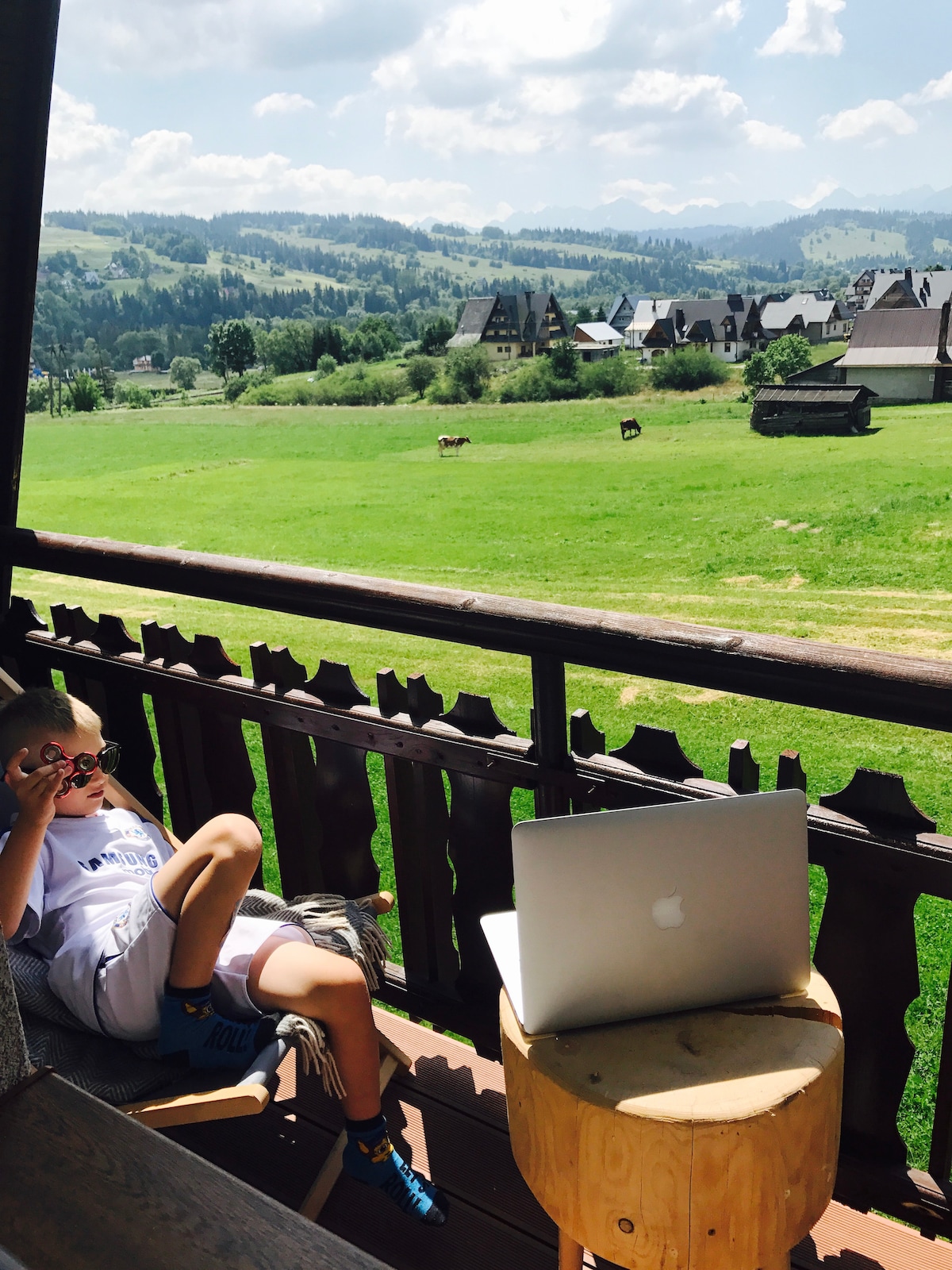 A balcony setup is depicted with a wooden stool and an open laptop positioned on it. A child is seen relaxing against the balcony railing, overlooking a lush green landscape with distant houses and rolling hills under a bright sky.