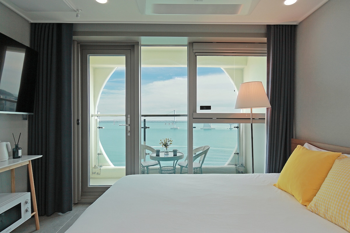 A spacious room with large windows offers a view of the ocean and a balcony. A white bed with yellow accent pillows is positioned in front of the windows. A small table and two chairs are visible on the balcony, providing a relaxing space to enjoy the scenery.