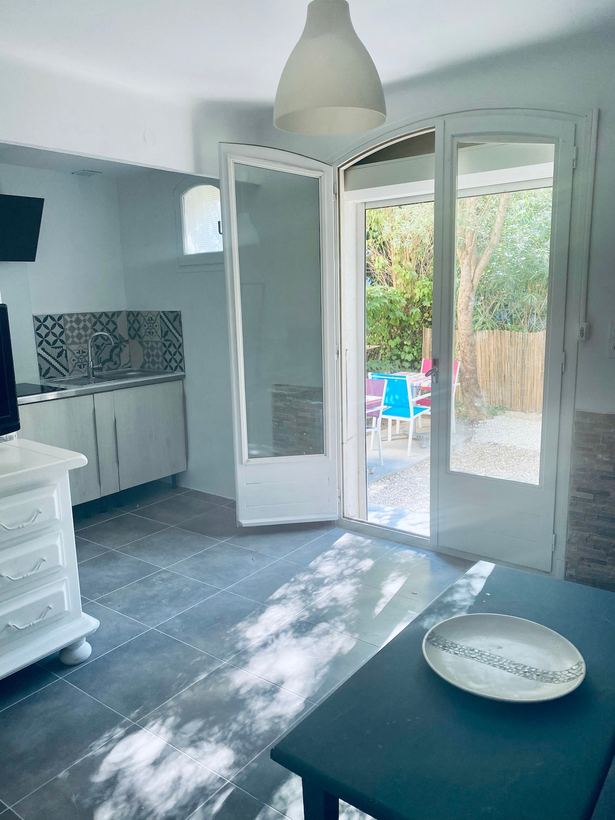 A light-filled interior is shown, featuring large glass doors that open to a private garden. A modest dining area is visible, with a table and chairs ready for use. The space is enhanced by neutral tiles and minimalistic decor, creating a calm atmosphere.
