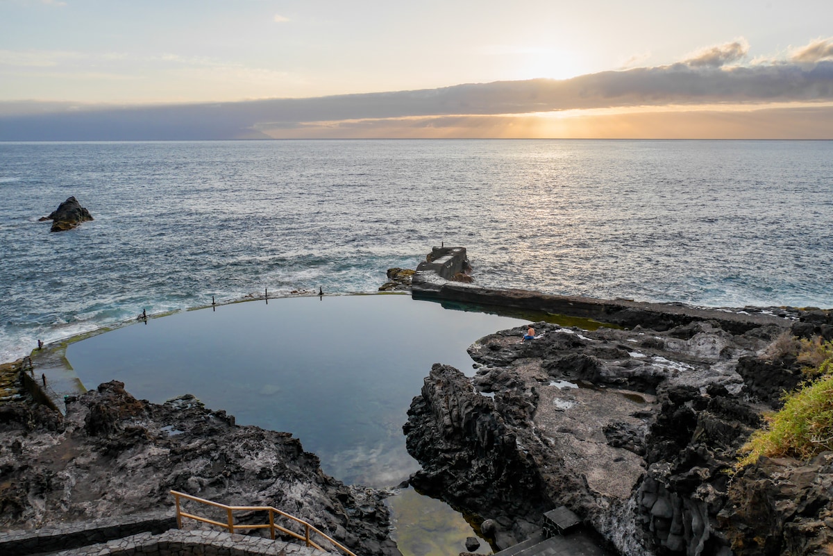 A natural swimming area is framed by rocky formations and ocean views under a sunset sky. The calm water reflects the light of the setting sun, while people can be seen enjoying the space along its edges.
