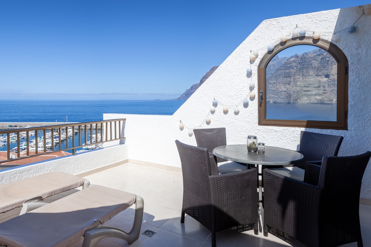 A terrace features a round table surrounded by four dark wicker chairs, offering a view of the clear blue ocean and nearby cliffs. A sun lounger is positioned to the side, and decorative string lights enhance the space. A large window captures the panoramic views.