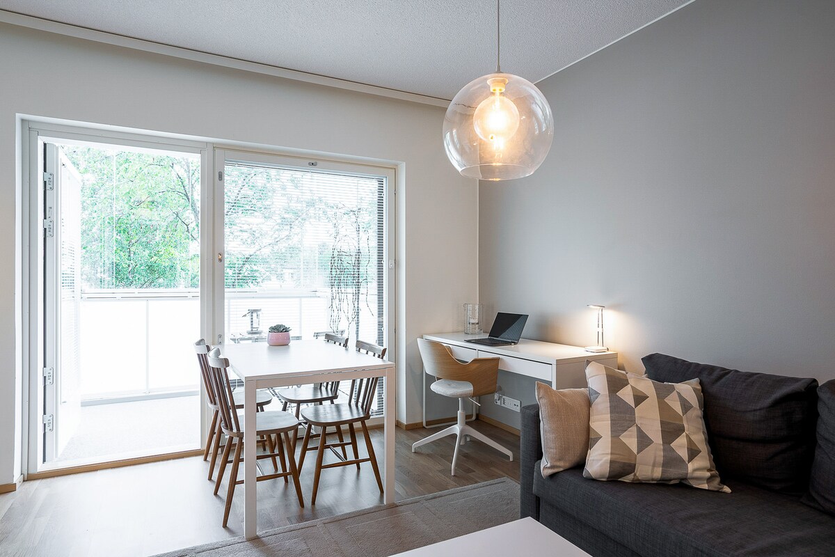 A bright living area features a comfortable sofa and a light-colored dining table with chairs. A glass light fixture hangs above the table, while large windows provide a view of the balcony and surrounding greenery.