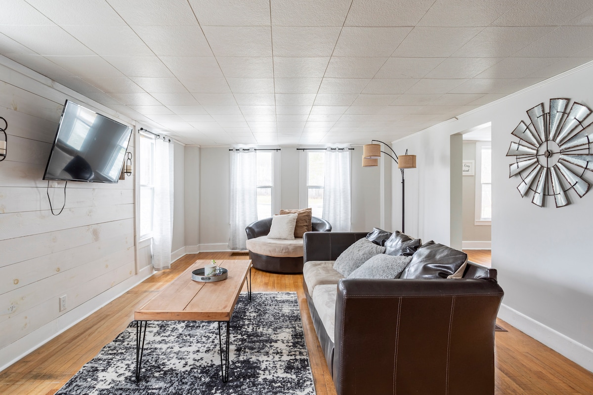 A bright living area features a large sofa and a matching armchair, with a wooden coffee table at the center. Natural light filters through sheer curtains, and a television is mounted on the wall. A decorative mirror adds a touch of elegance to the space.