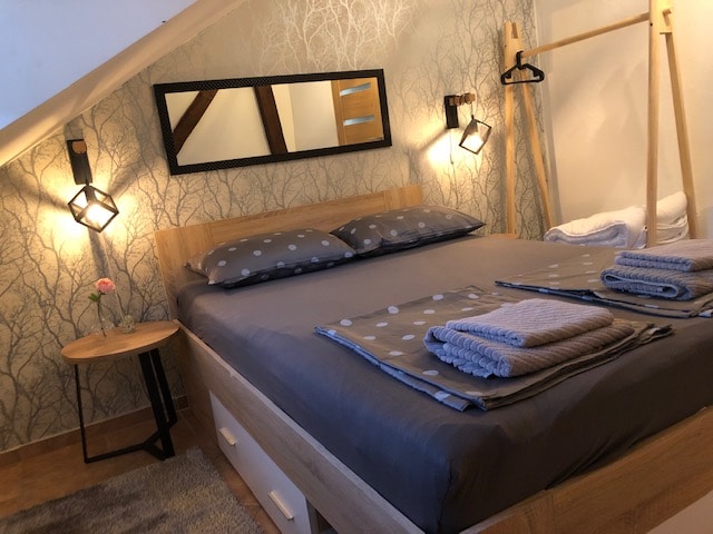 A well-appointed sleeping area features a spacious bed with dotted bedding. A wooden side table is situated next to the bed, with a small flower vase atop. Soft lighting comes from wall sconces, and a stylish mirror is mounted on the wall, enhancing the decor.
