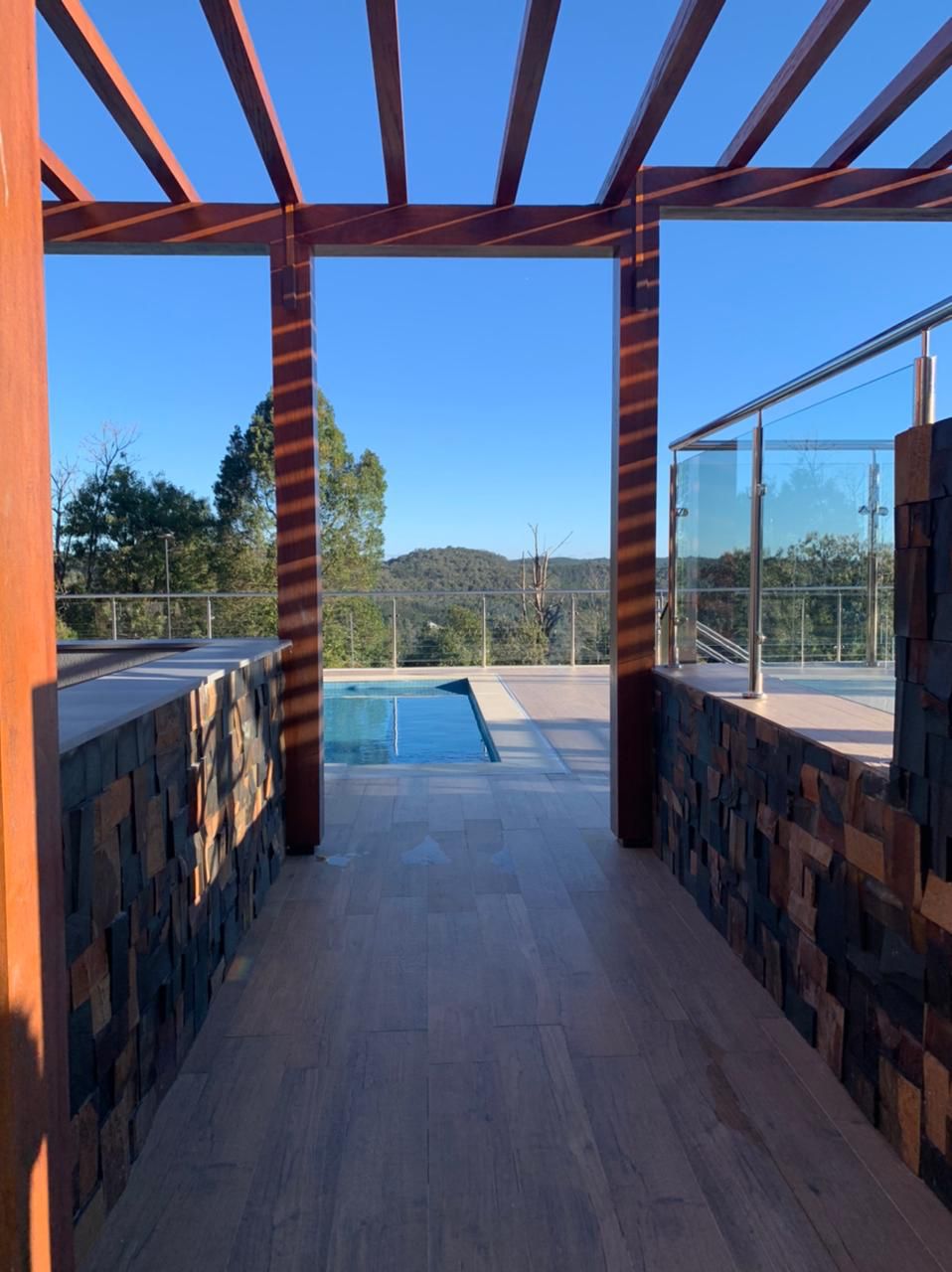 A pathway framed by wooden beams leads to a clear pool, surrounded by a glass railing. The view includes distant hills under a bright blue sky, showcasing nature's serene landscape.