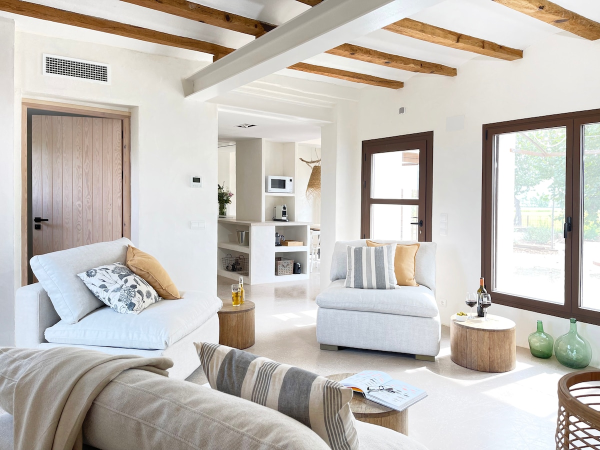 A spacious living area features light-colored walls and wooden beams across the ceiling. Two comfortable sofas are arranged, alongside a round wooden table. Natural light fills the space through large windows, enhancing the open layout that connects to a kitchen area in the background.