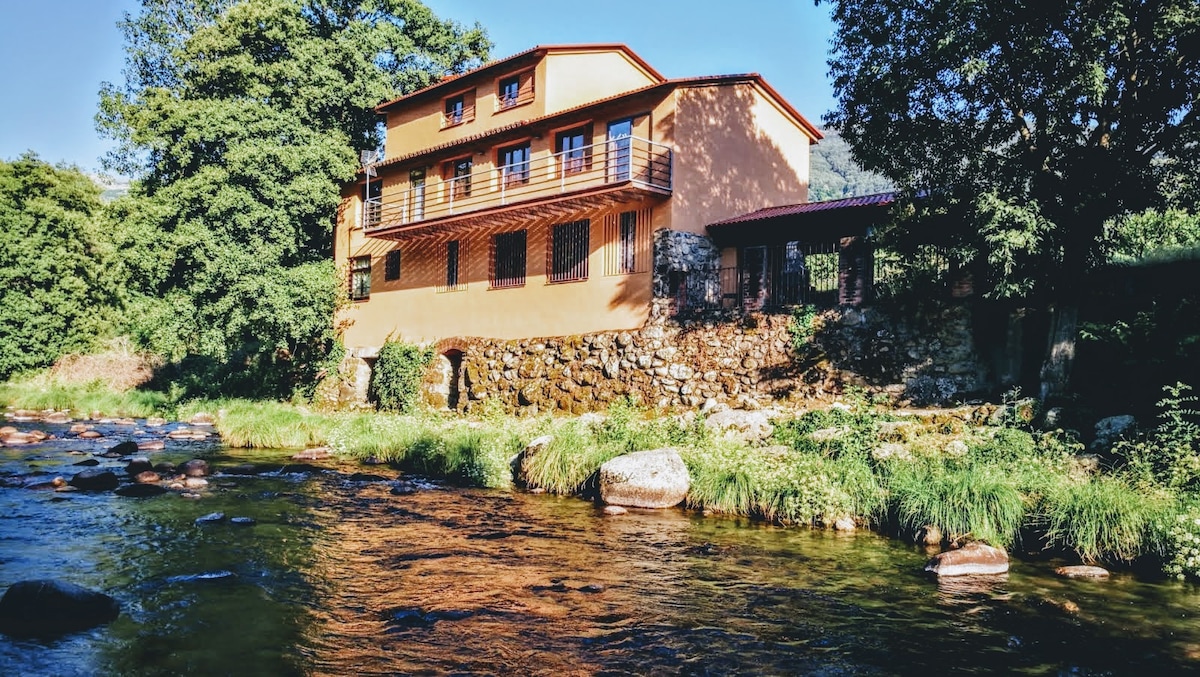 The rustic building, nestled by the riverbank, features a warm orange facade with multiple windows. Lush greenery surrounds the property, and smooth stones lead into the gently flowing water, reflecting the building's inviting atmosphere.