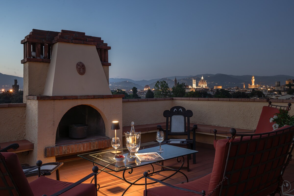 A spacious terrace is illuminated softly in the evening, featuring a fireplace and comfortable seating. A glass table holds various items, while the stunning skyline of Florence, including its notable towers and landmarks, is visible in the background against a twilight sky.