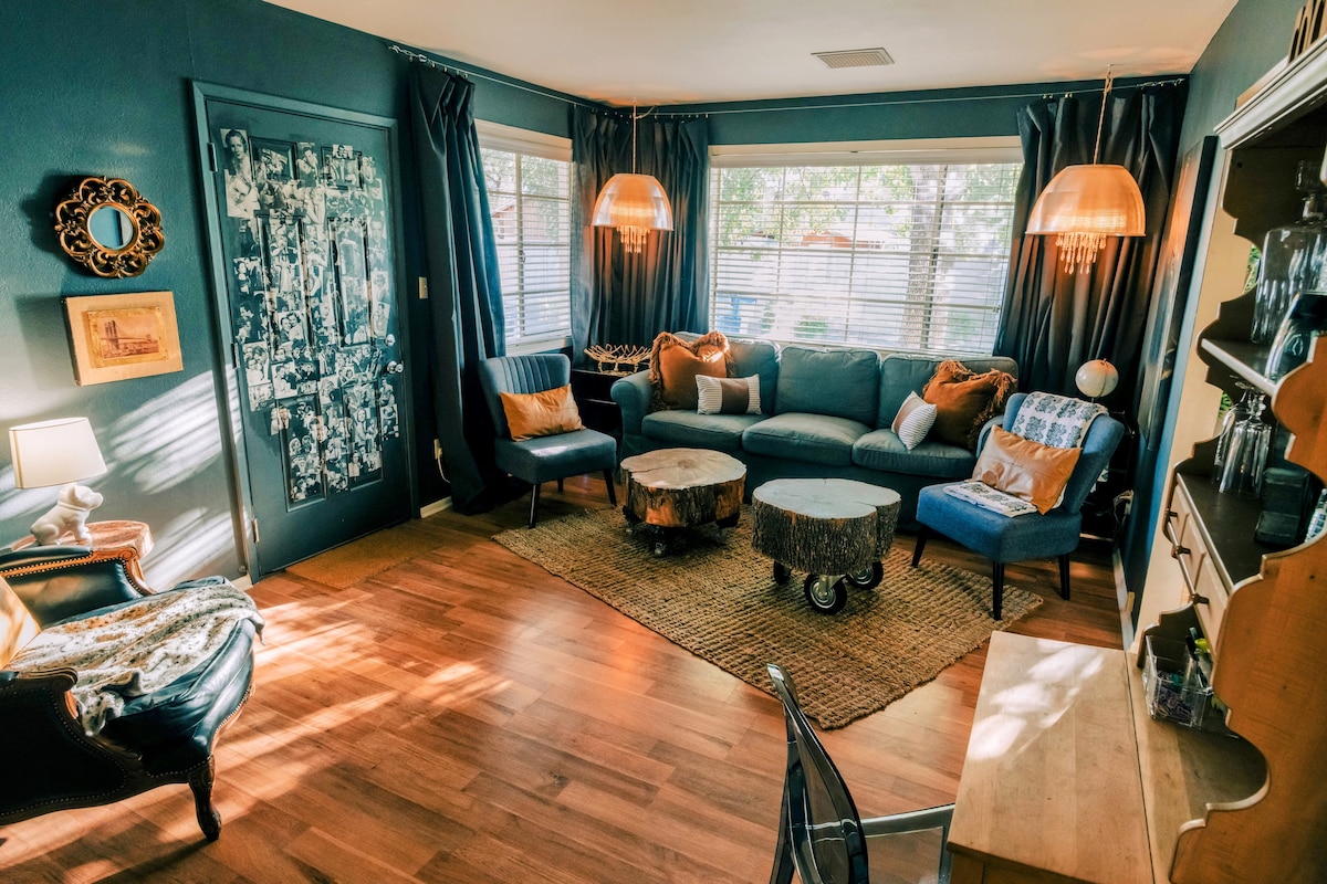 A cozy living room is shown, featuring a comfortable couch, two side chairs, and a large vintage lounge chair. Natural light streams through the windows, illuminating the wood flooring. Unique pendant lights hang above a custom coffee table made from a tree stump.